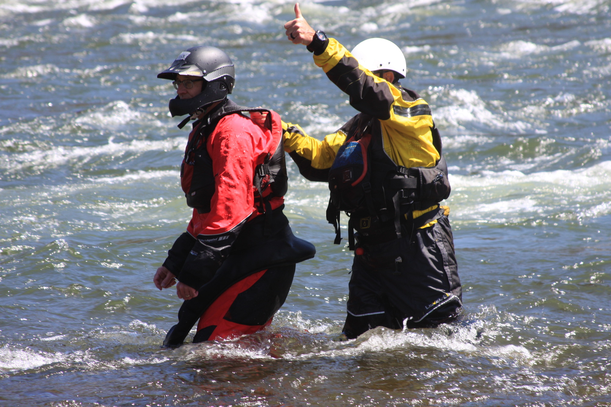 Two people in life jackets and helmets in knee deep water in the river. One person gives a thumbs up.
