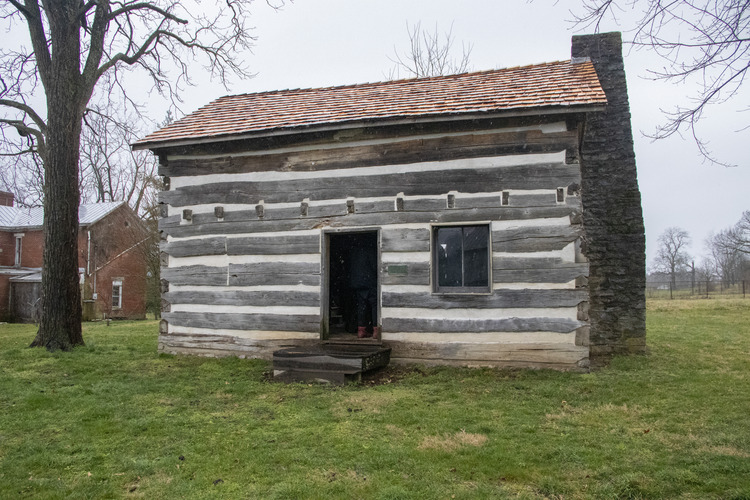 A wooden cabin with white mortar between logs and a red shingle roof and stone chimney to the right.