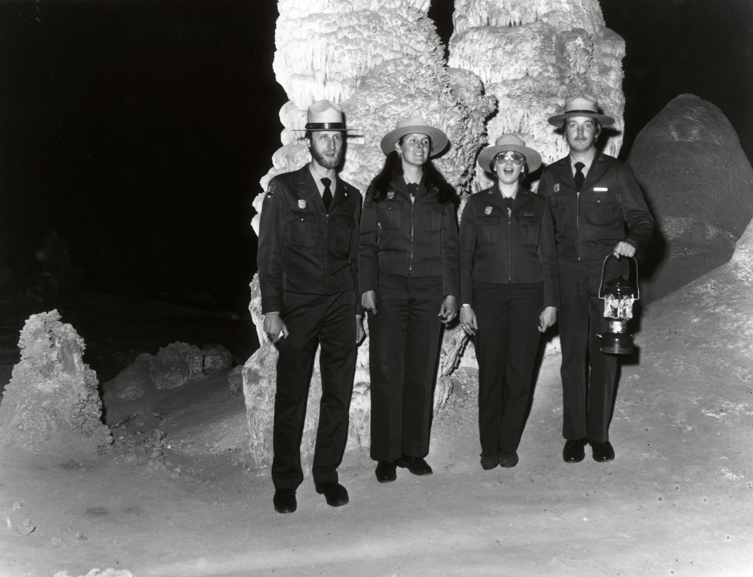 Two women and two men rangers in NPS uniforms and broad-brimmed hats stand side by side inside a cave. One ranger holds a lamp. 
