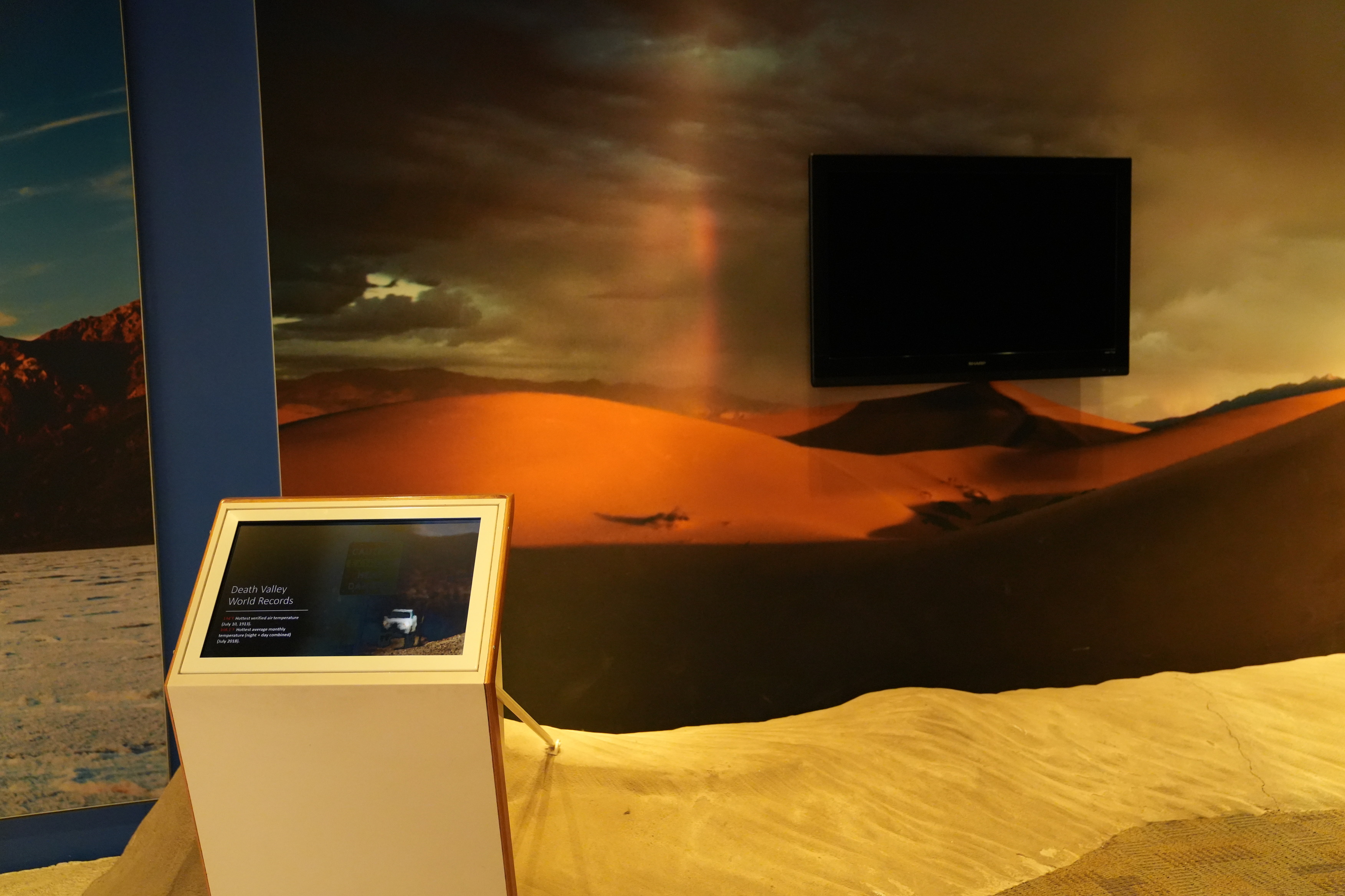 Furnace Creek Visitor Center Museum Why is Death Valley so Hot exhibit. A screen at waist level is shown displaying weather facts about Death Valley. A blank TV screen in on the wall. This typically plays an informational video. Tactile, textured sand dunes boarder the area between the wall and the waist-level screen.