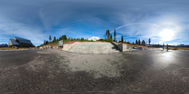 A set of stone steps with inscribed with the text "...the most luxuriant and the most extravagantly beautiful of all the alpine gardens I ever beheld in all my mountain-top wanderings. - John Muir, conservationist, 1889" below a view of Mount Rainier surrounded by meadows, paved pathways, and a large building with slated roof. 