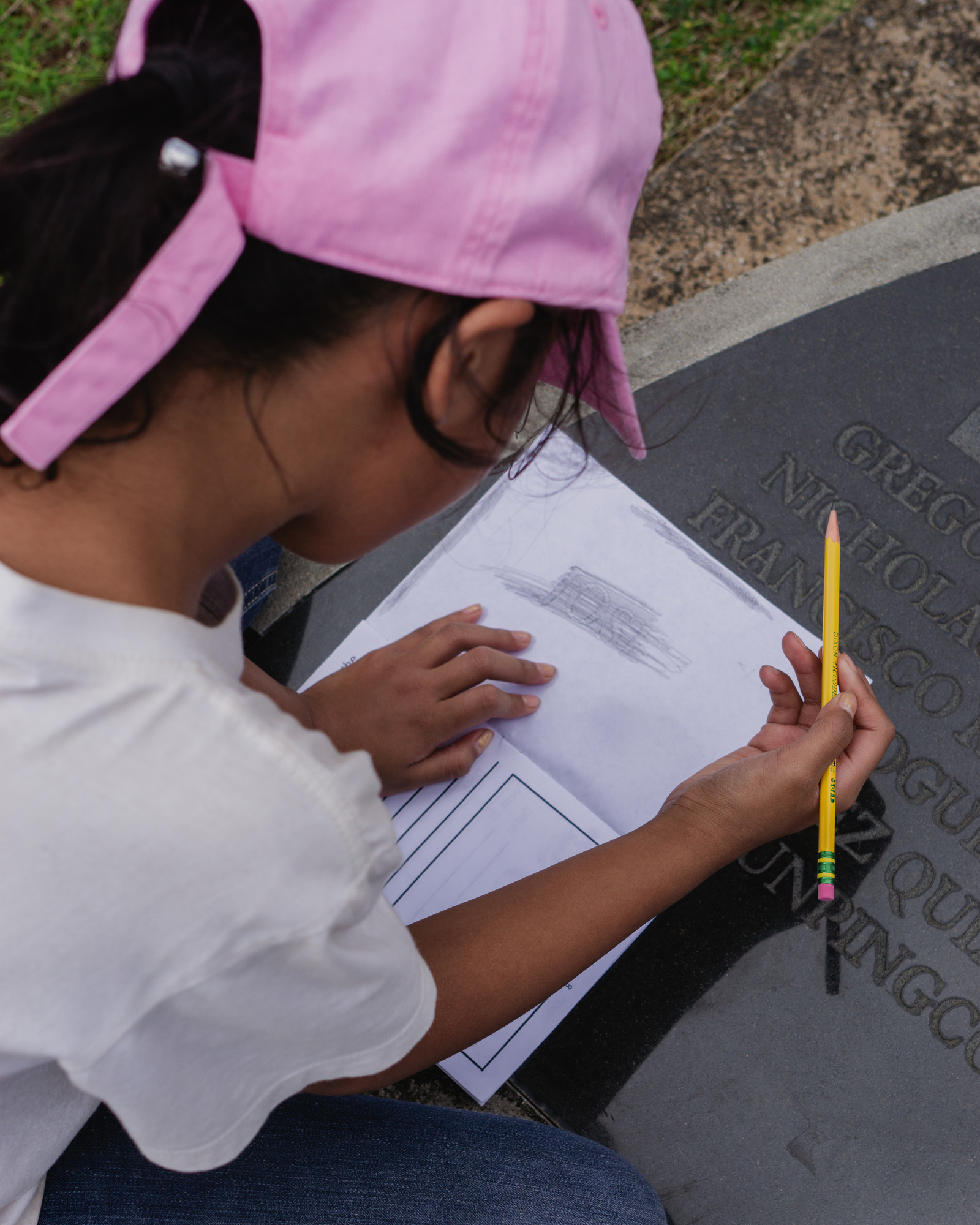 A girl in a pink hat makes a rubbing of a name engraved on a plaque.