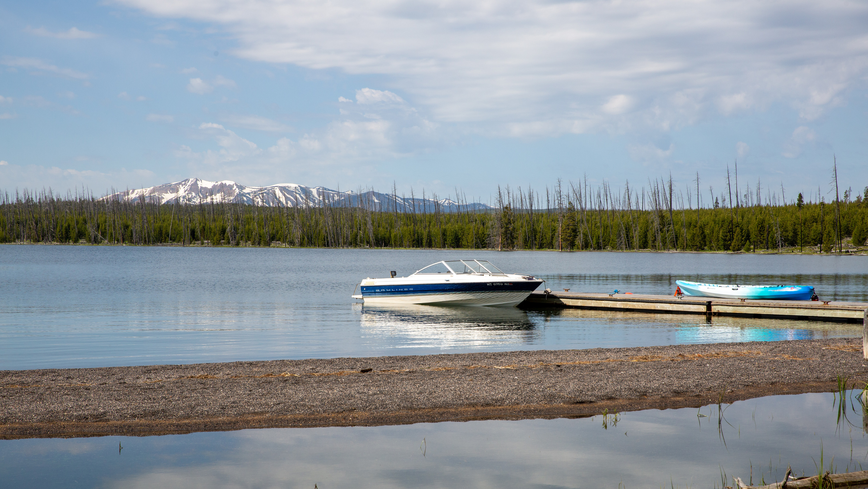 A small motorboat and a blue kayak are both tied up to a dock on a lake with a snow covered mountain in the background
