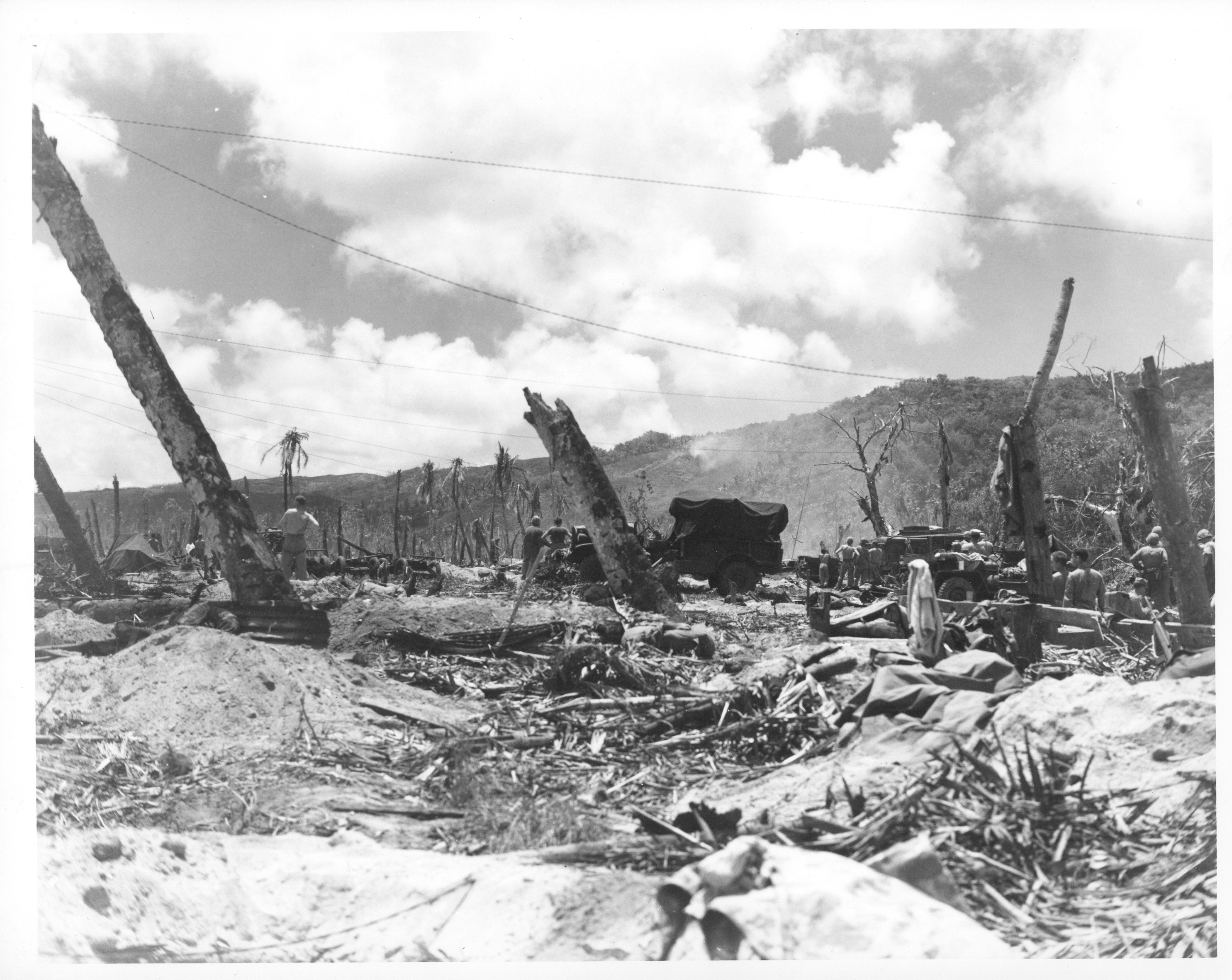 A beach full of craters, burnt and twisted trees and destroyed buildings and vehicles.