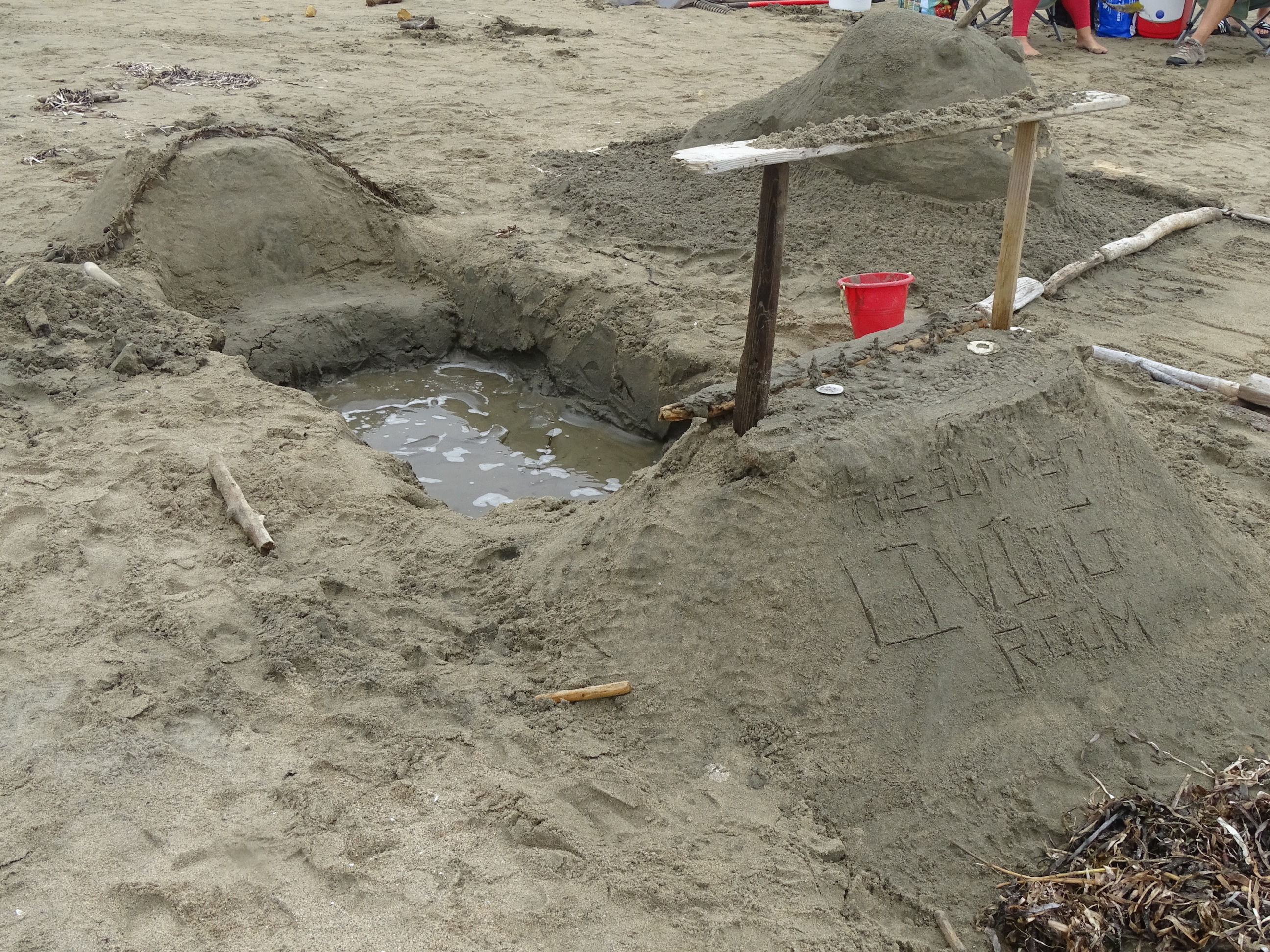 A sand sculpture of a water-filled hole with two mounds of sand on both sides. The mound on the right is topped by an arch made of driftwood and the words "The Sunken Living Room" written in the mound.