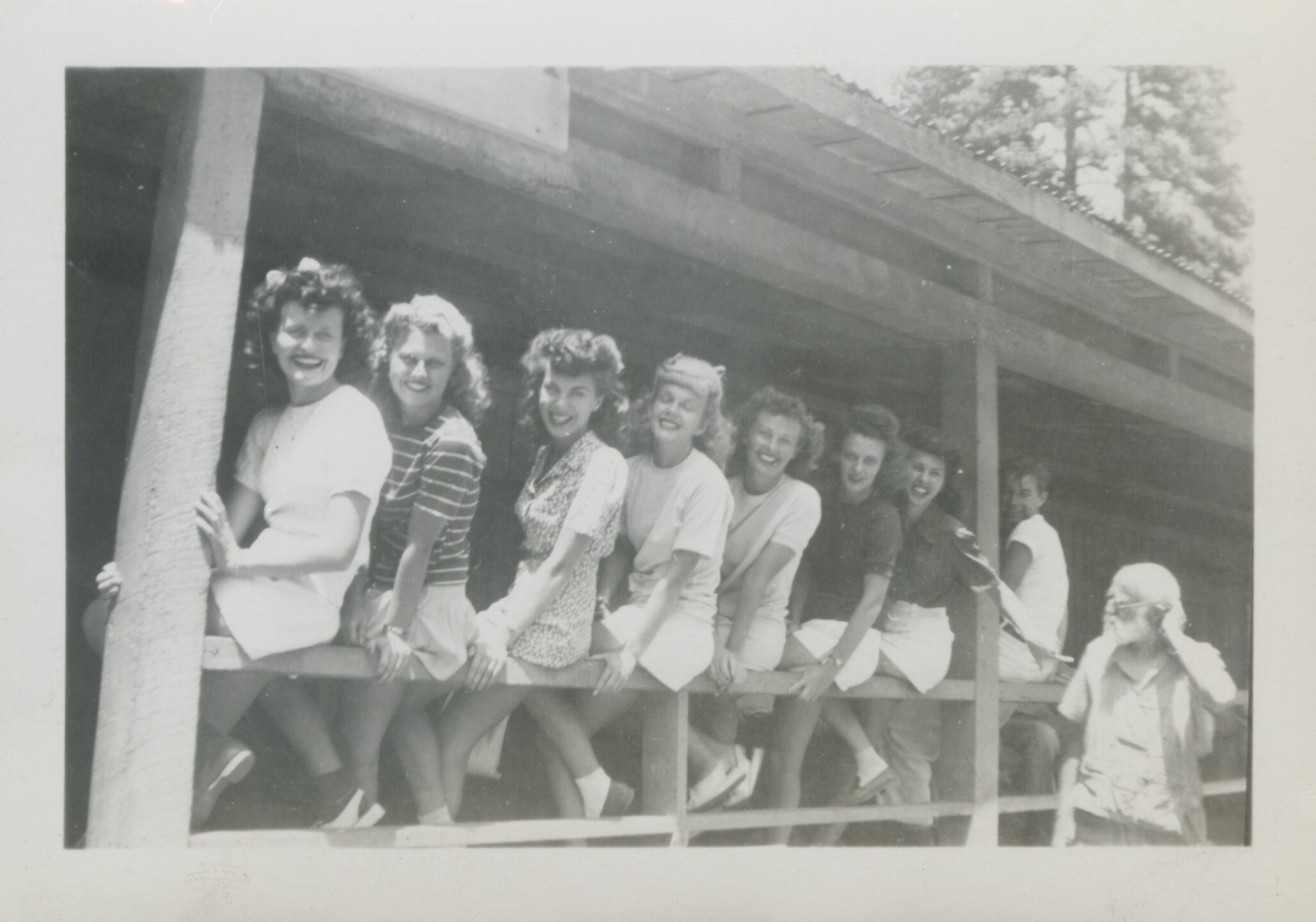 Seven nurses pose on a porch railing, also two unidentified males in the shot
