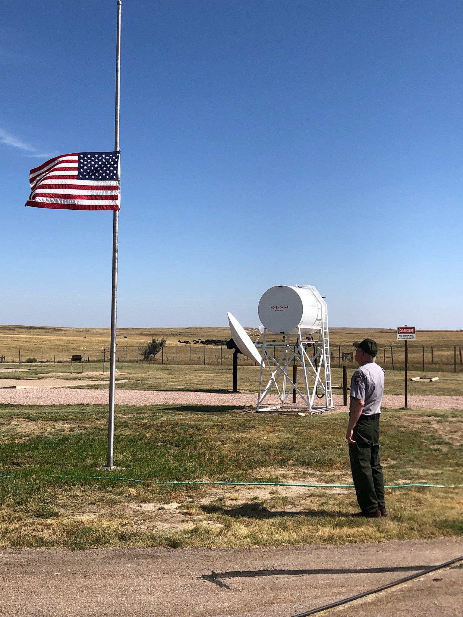Man in a park service uniform of gray shirt and green pants saluting the US flag flying at half mast. 
