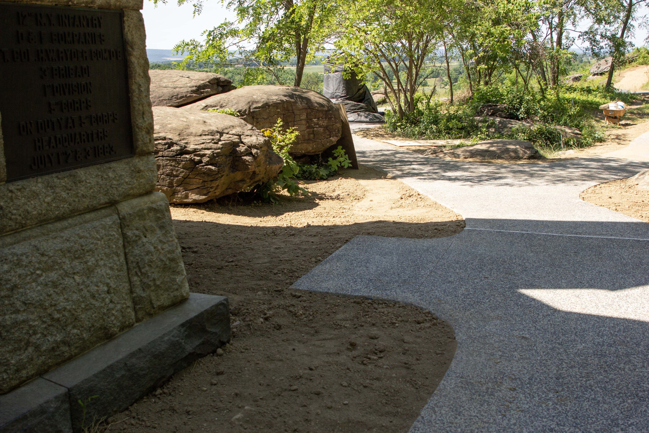A concrete walkway sits to the right of a large stone monument and runs to the left between boulders and trees and to the right beside a patch of trees.
