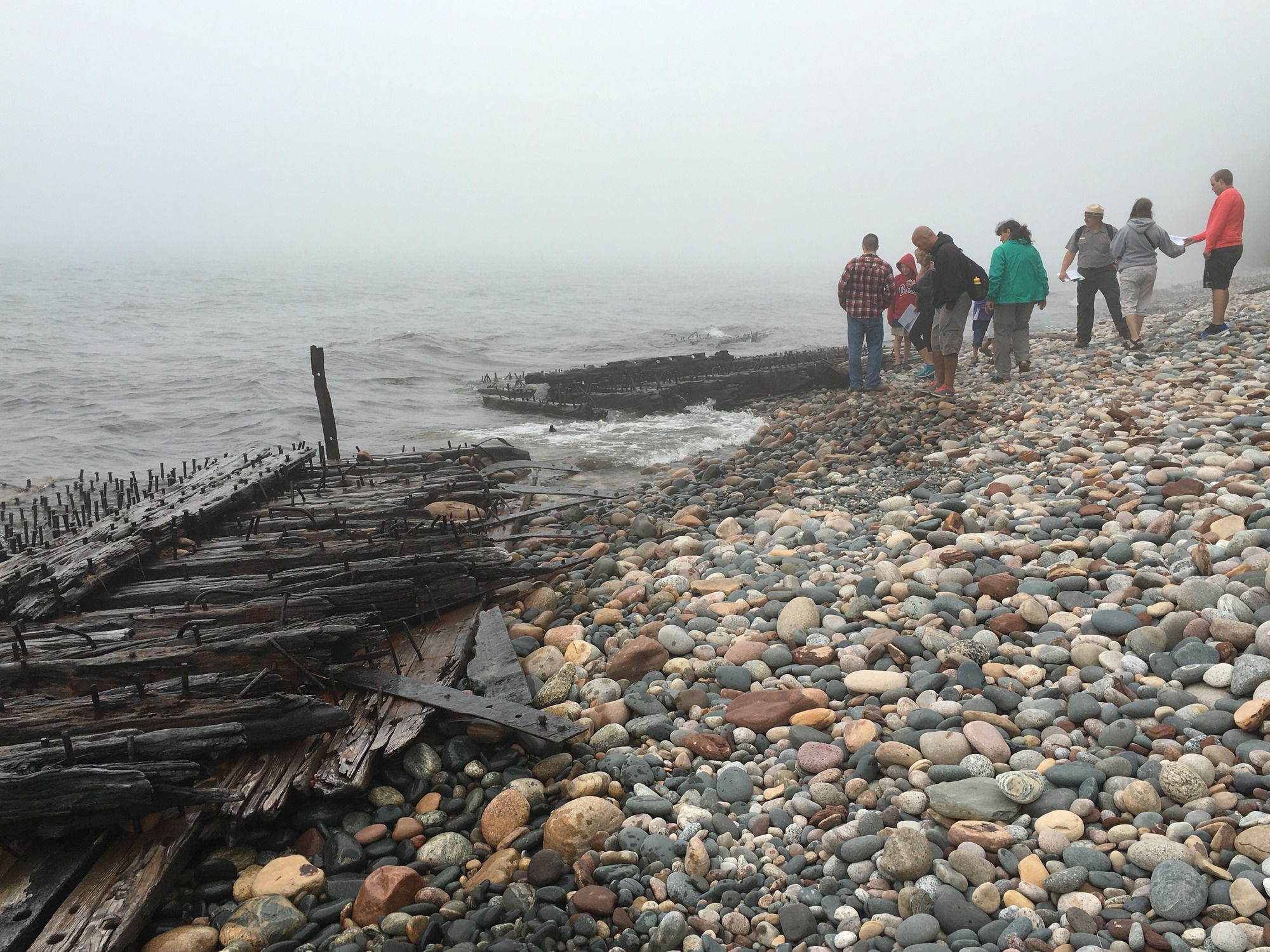 Park ranger and group of people on a rocky Lake Superior beach looking at wooden parts of old ships that washed ashore. The ship pieces are made up of wooden boards, metal straps, and metal studs. It's a foggy day.