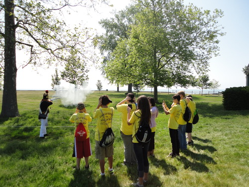 A man dressed as a US Marine from the war of 1812 fires a musket away from a school group of about a dozen people. The students and adults are either are taking pictures with their phones or covering their ears.
