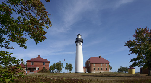 A tall white and black lighthouse surrounded by red brick buildings and trees that are just beginning to turn red. 
