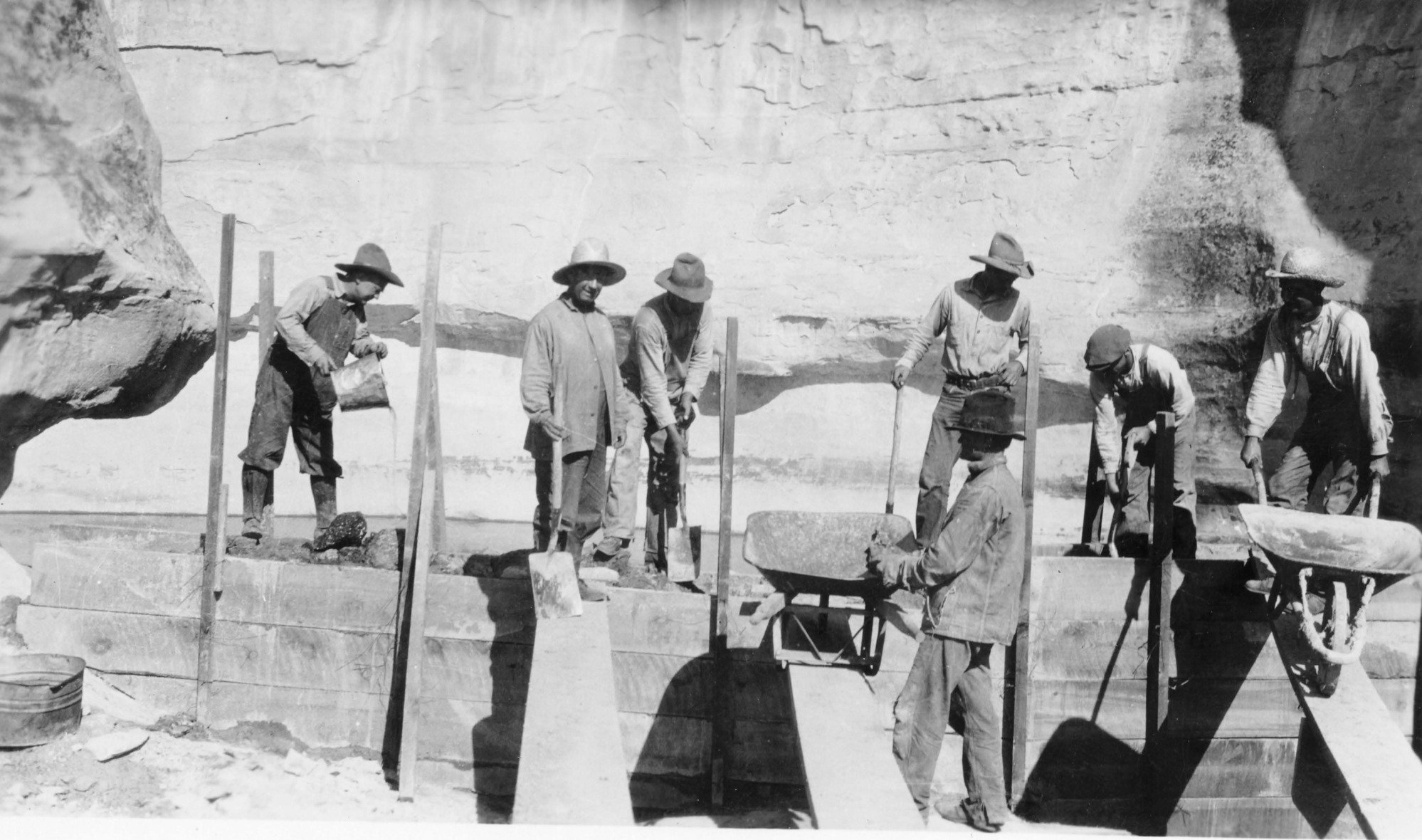 A black-and-white photo of the construction of the dam around the pool of El Morro. Seven men work with shovels, buckets, and wheelbarrows around a wooden frame.
