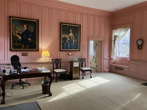 A plantation house entrance hall with pink painted wood paneling, two impressive portraits of a man and woman in upper class 18th Century style, a desk, and a donation box.