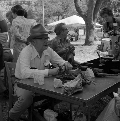 Bill Miller carving a peach pit creature at the first annual Folklife Festival, Zion National Park Nature Center, September 1977. Leo Fesler talking with visitor in background
