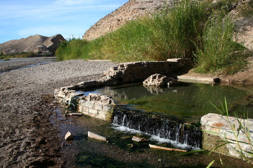 A pool of water pours out of the ruined foundation of a desert hot spring bath house.