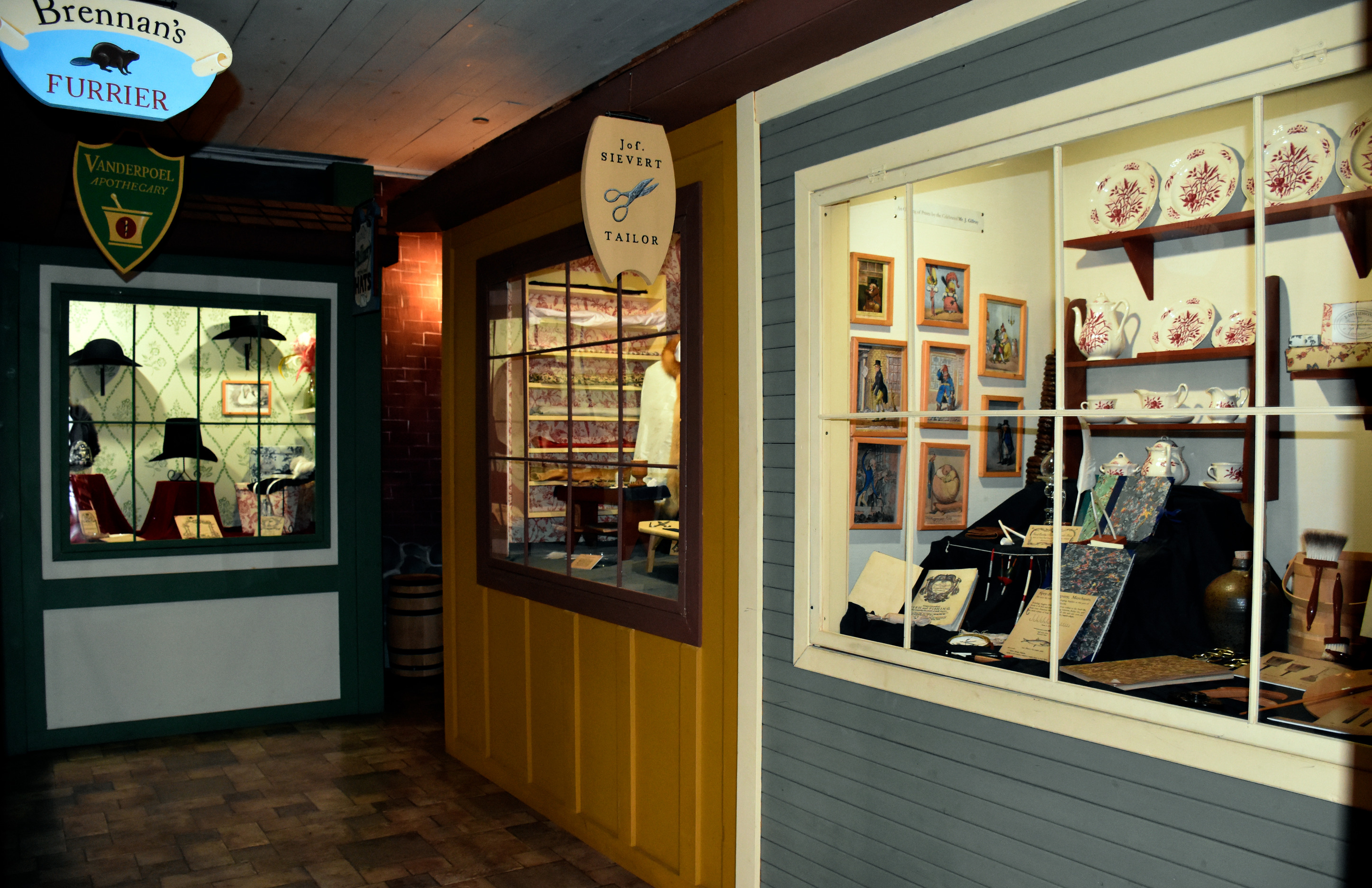 A dark corner with three illuminated windows. One shows dishes, fancy books, and small illustrations. Another shows fabrics, and the third various historic hats.
