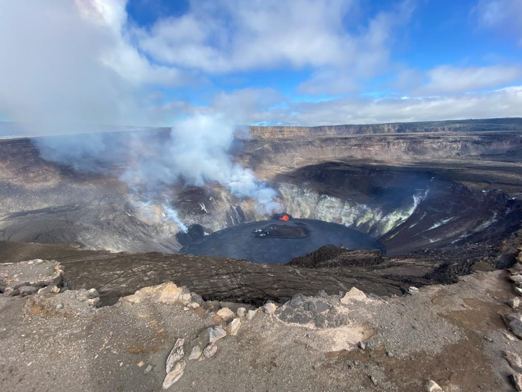 Black lava lake in a volcanic crater sending out a white plume of steam beneath blue skies