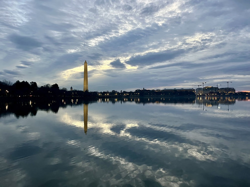 Large tidal basin lined with trees with the reflection of the trees, Washington Monument, and clouds reflected in the water under an early morning sky