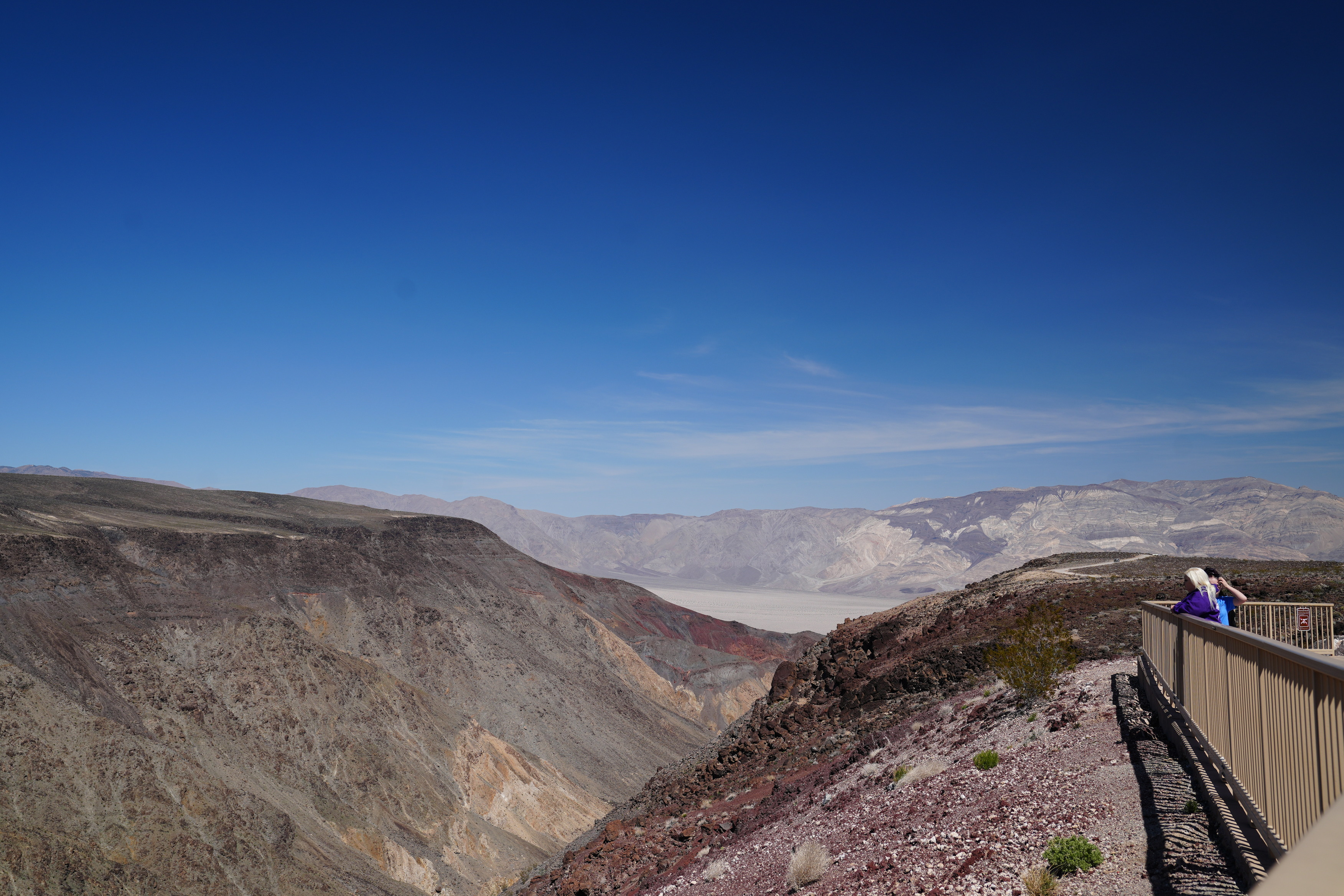 Father Crowley's Vista viewpoint. A railing surrounds the sidewalk zone where two visitors are overlooking the canyon. The canyon is rainbow in hue. 