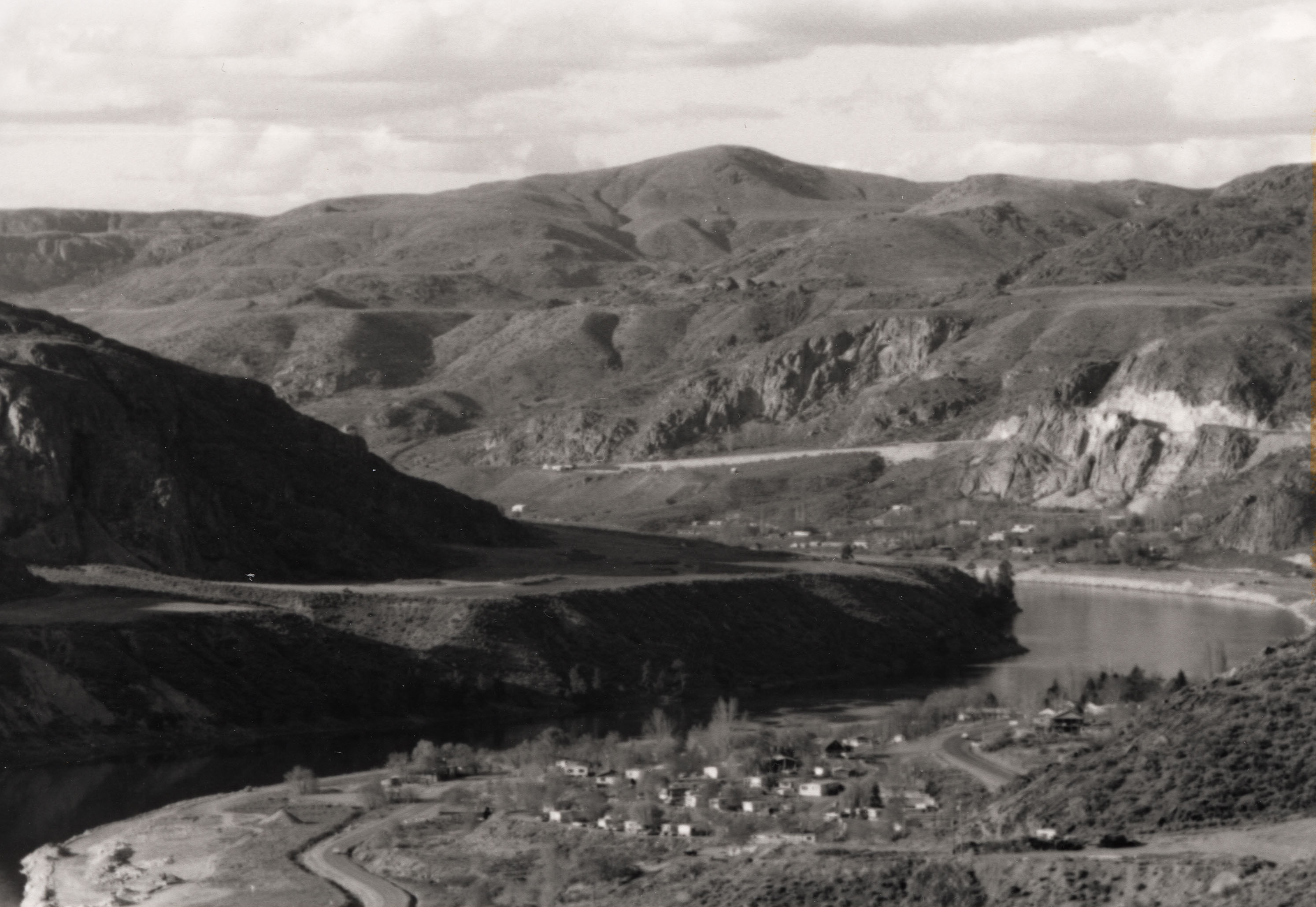 Black and white photograph of small town next to a river bend with bare mountains rising in the background