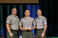 A man holds an award with two men in gray and green uniforms at an award ceremony.