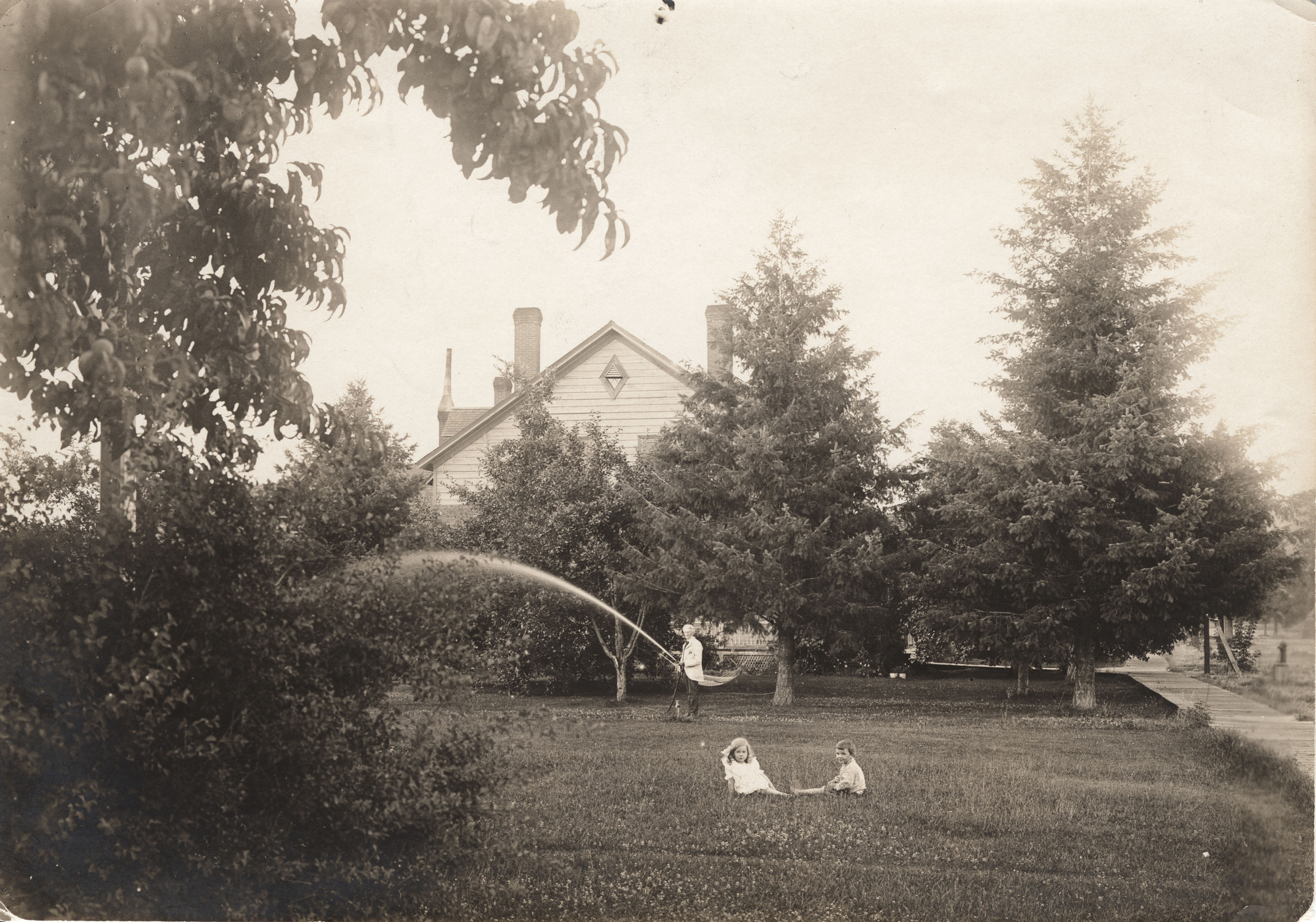 Black and white photograph of two young children sitting on a lawn while a man waters the grass behind them