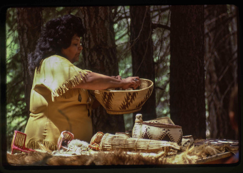 A woman in a Native American attire holds a woven basket above a table of Atsugewi crafts. 