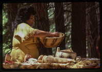 A woman in a Native American attire holds a woven basket above a table of Atsugewi crafts. 