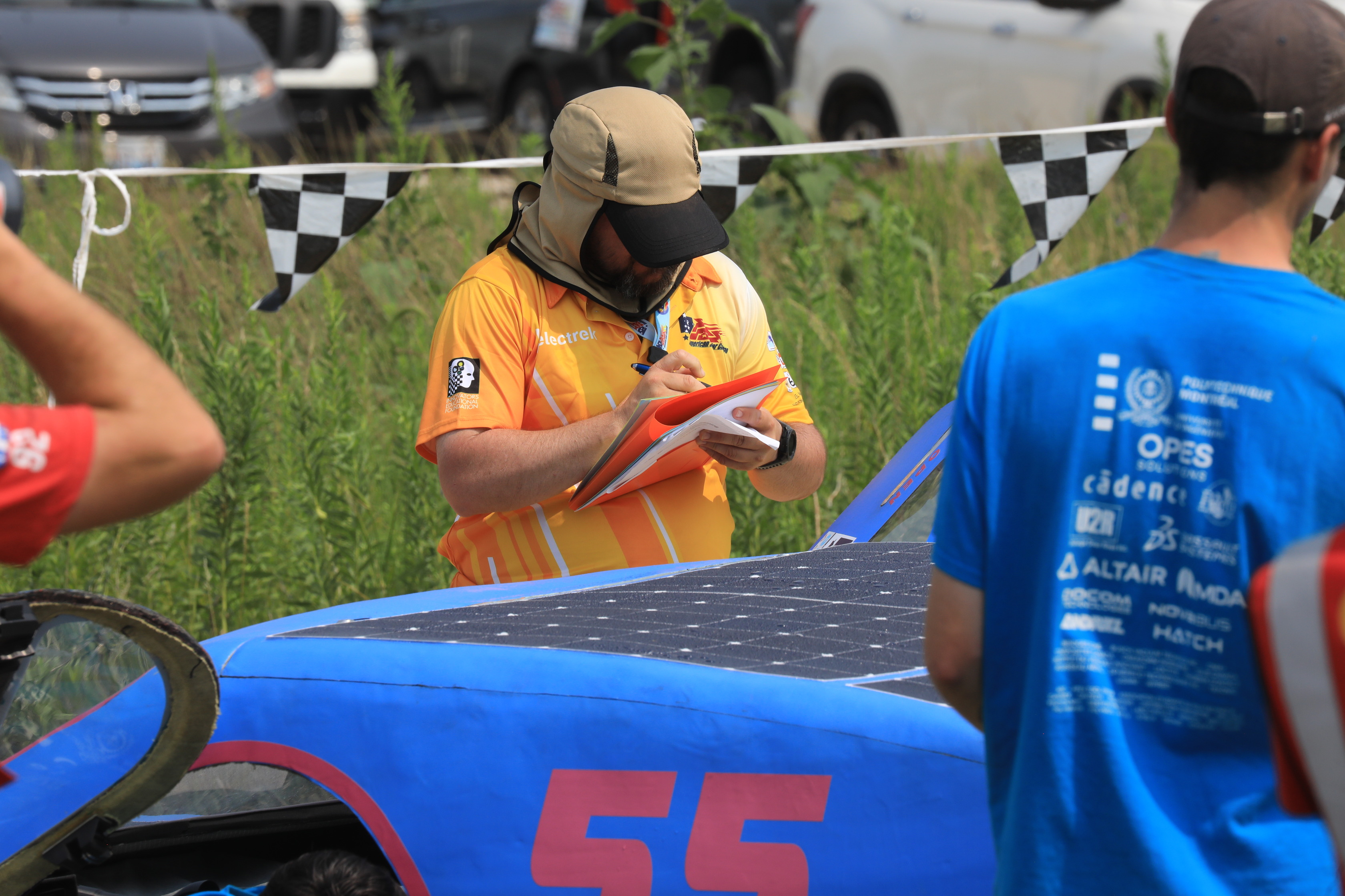 Man with orange-yellow shirt takes notes while looking at a blue and pink solar car. Small beads of water can be seen across the solar panels.