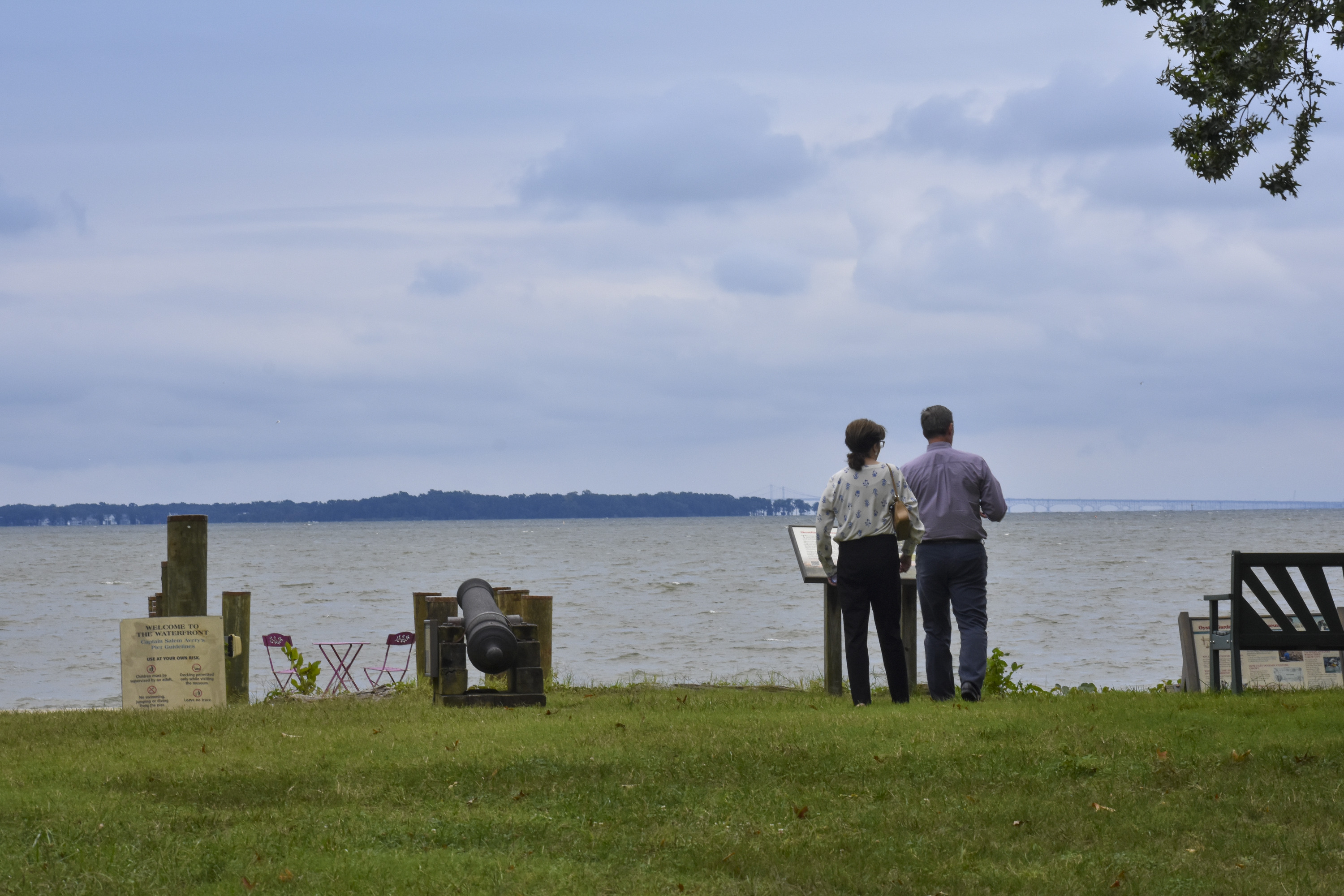 Two people stand by a waterfront with an informational sign, overlooking a body of water and distant bridge.