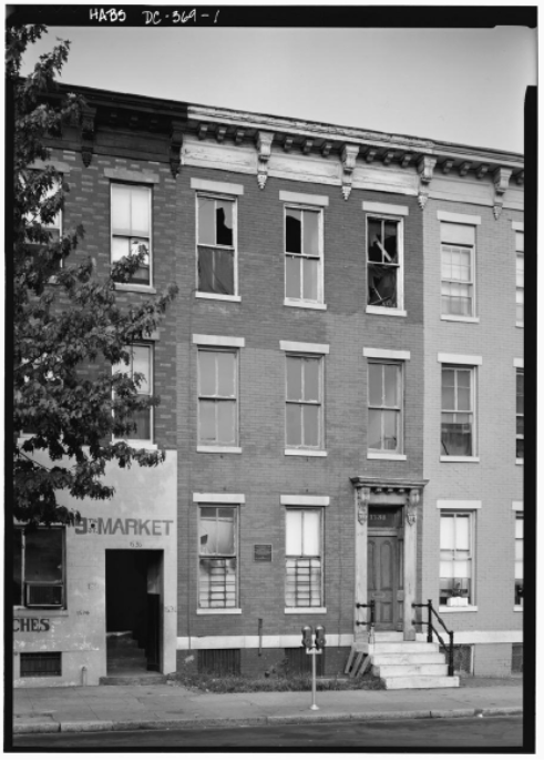 A brick row house shown from the street view. Several windows are broken and boarded up. It sits between two other row houses.