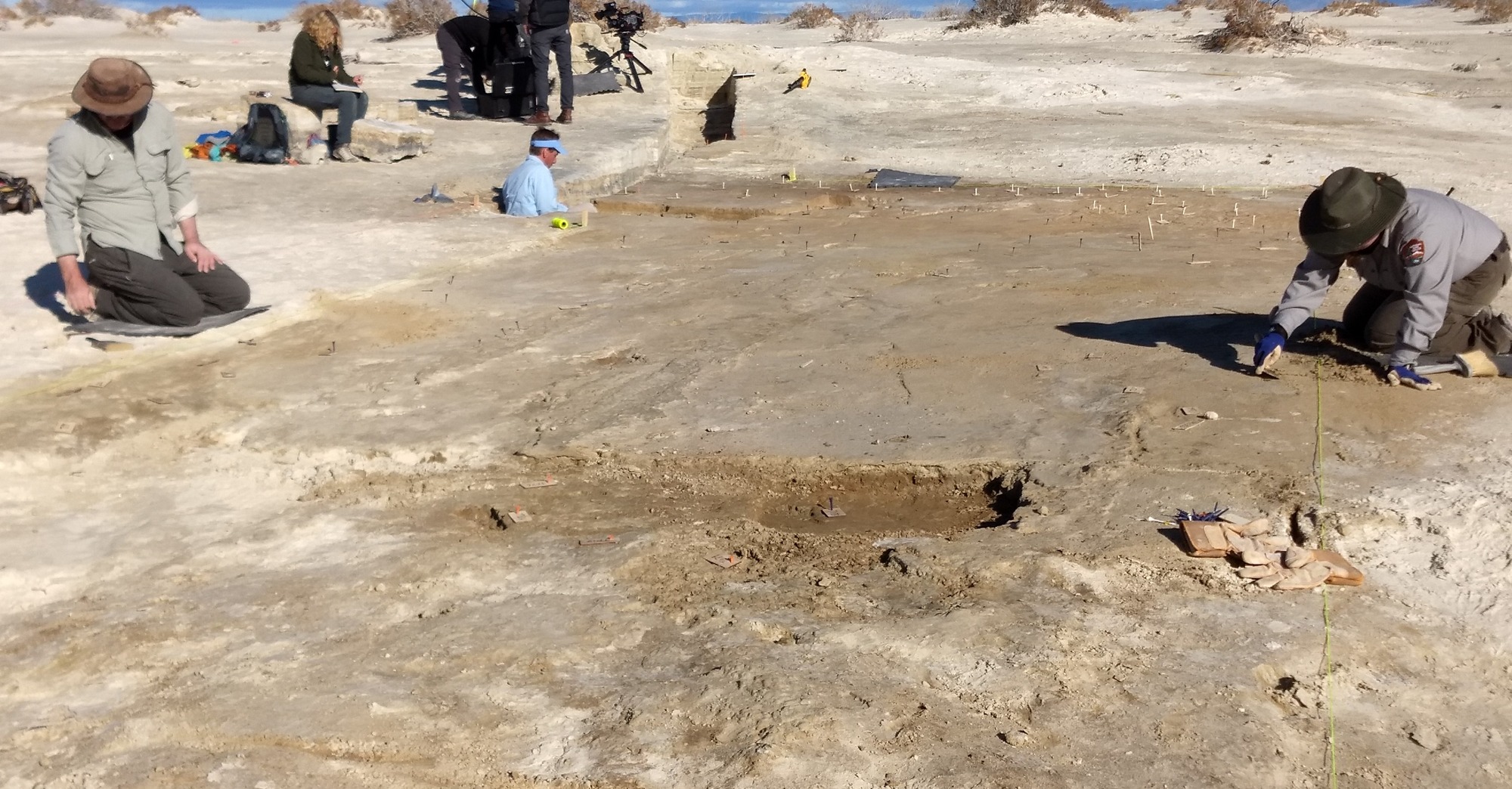 Multiple researchers kneeling over a site plot and using tools to reveal the footprints fossilized in the gypsum sand. 