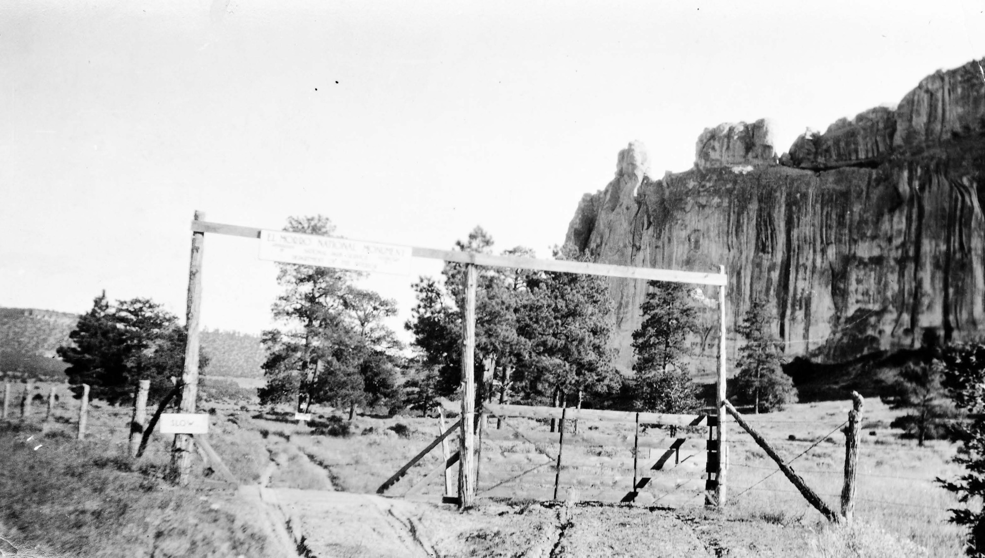 A black-and-white photo of a fence line. Tall poles hold a sign overhead that reads "El Morro National Monument." A dirt road leads underneath. El Morro's rock face looms in the background. 