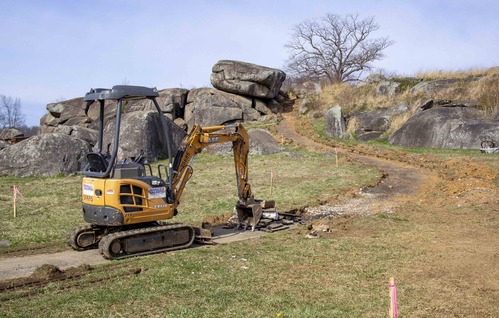 In the center-left is an unmanned yellow front-end loader on a dirt road. There is a pile of concrete chunks in front of the vehicle’s claw in the road. The road curves up from the left to the center background.
