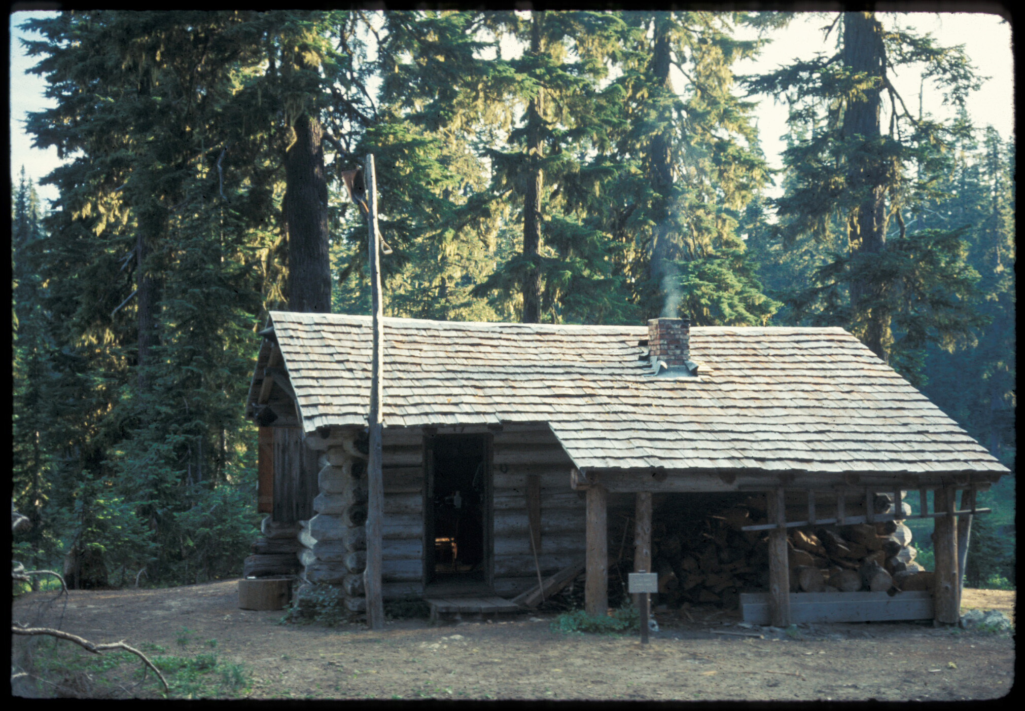 A log cabin with a covered porch in front of half the building with a fireplace and flagpole. 