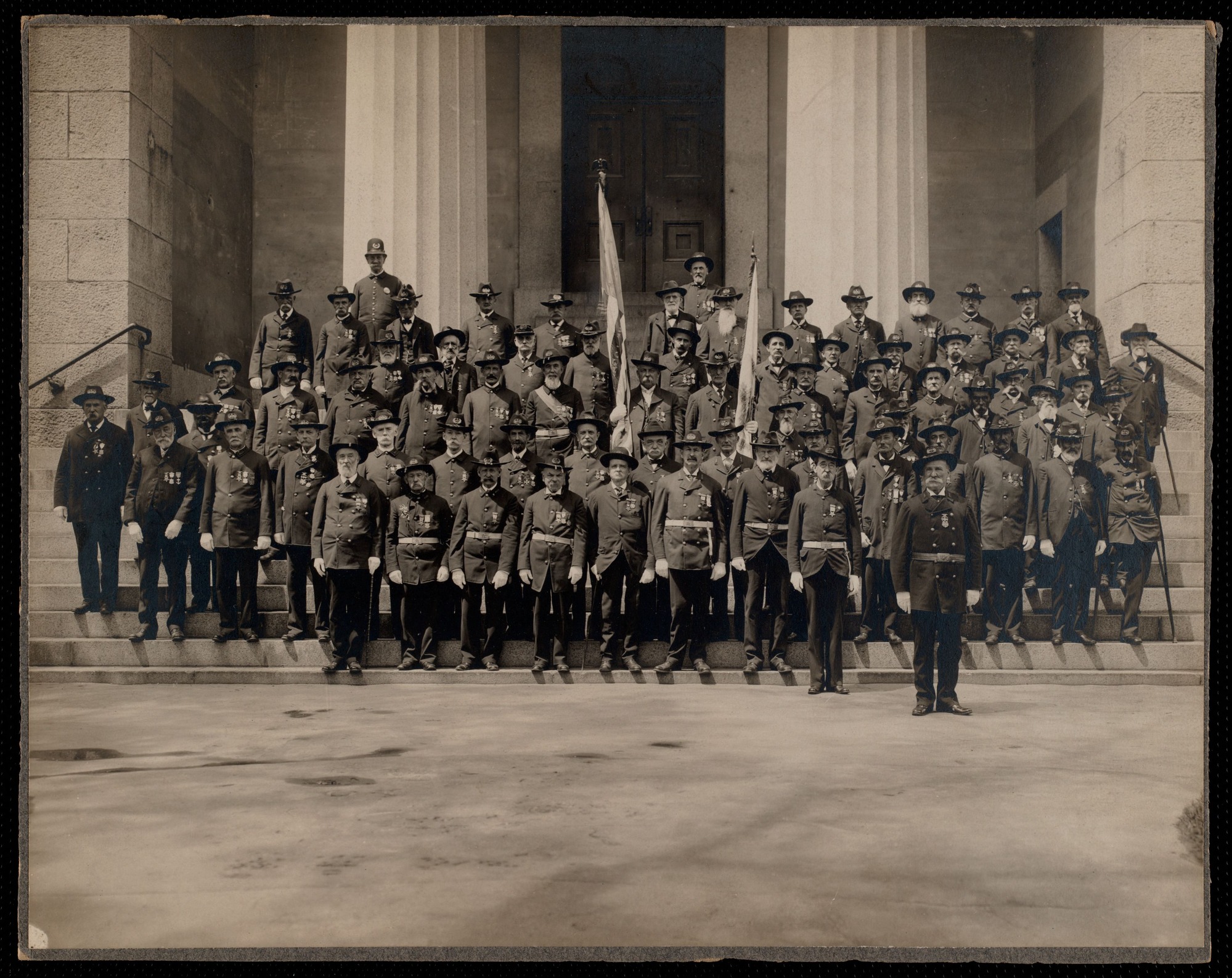 Black and white photograph of members of the Grand Army of the Republic Post 1 standing in front of a building with white columns. All men are dressed in uniform.