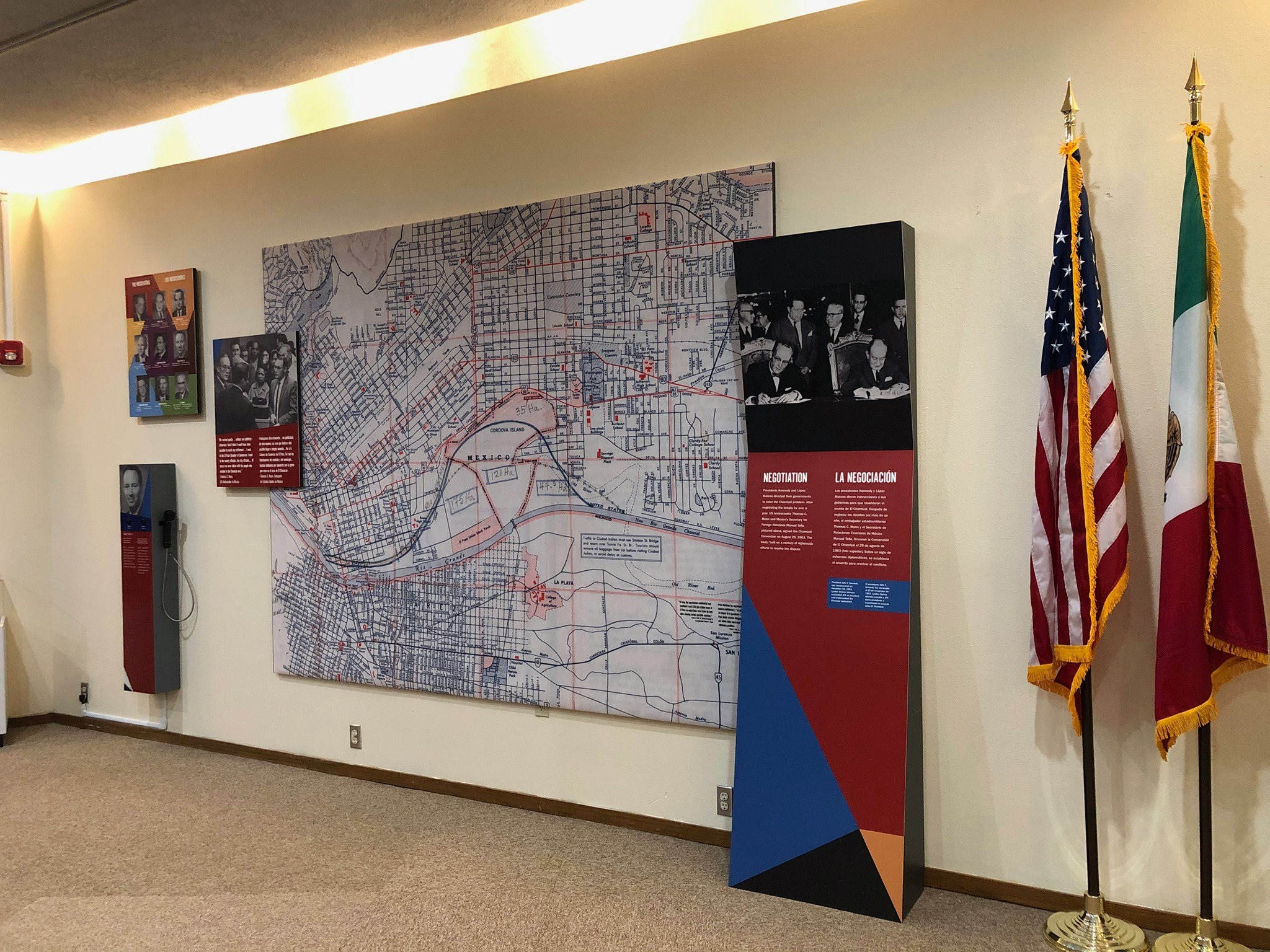 Exhibit panels and a listening station are fixed to an indoor wall. US and Mexican flags stand on the floor to the right side.