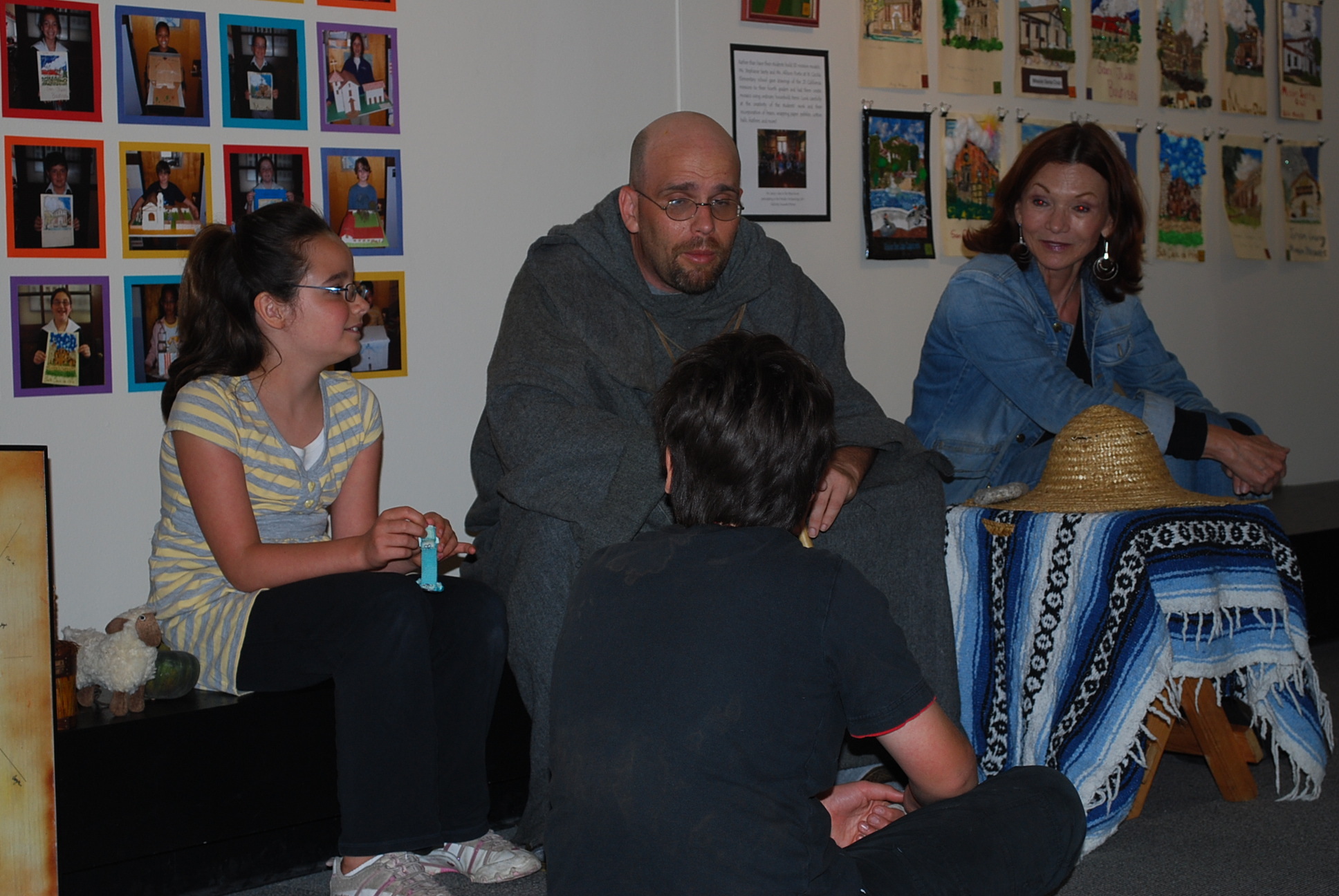 A bald man with a beard and glasses speaks to an older woman with long brown hair and two kids