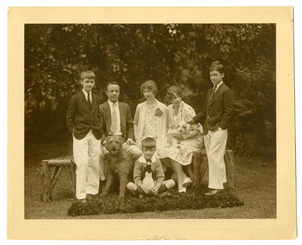 A man, his wife, and teenaged daughter sit on a wooden bench for a pose portrait outside. The daughter holds a puppy in her lap. Two young men stand on each side of the bench. A small boy sits at the feet of the mother next to a dog. The boy has a stuffed penguin in his lap.  