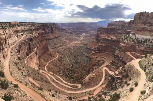 Steep, sandy switchbacks climb up a deep canyon. The sun is obscured by dense clouds. Mountains loom on the horizon.