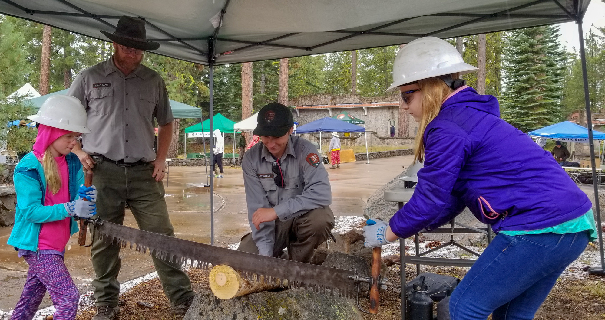 Two young girls hold either side of a long saw while cutting a small log. One ranger sits on the log and another stands and watches.
