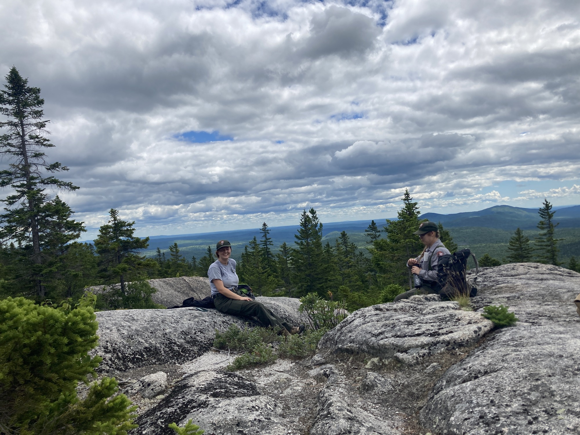 Two employees in NPS hats sitting on large boulders with a green landscape overlook.