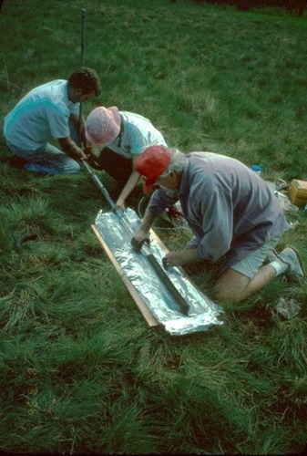 Three scientists kneeling in grass removing a sediment core from a metal tube to view it more closely