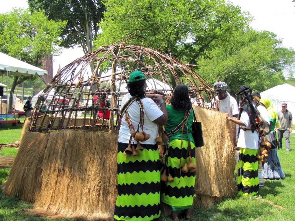 wooden dome frame has bottom half covered in grass, people in traditional dress are working on more