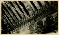 This is an historic black and white photograph from the Scotty's Castle Historic Photograph Collection, Death Valley National Park featuring detail of interior room ceiling. Elaborately cared wood beams parallel each other diagonally across face of image. A line of hexagon openings at junction of ceiling and wall overhanging front of stag where future medallions will be mounted. Below hexagon shapes is elaborate carved edge hanging down. Wood detailed stage ceiling seen bottom left. Decorative twisted rod upper right corner.