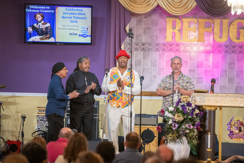 Bruce "Sunpie" Barnes and the Louisiana Sunspots can be seen in the center of the picture clapping while performing their last song of the night. Attendees are visible in the foreground looking towards the band. A purple and beige wall can be seen in the background.