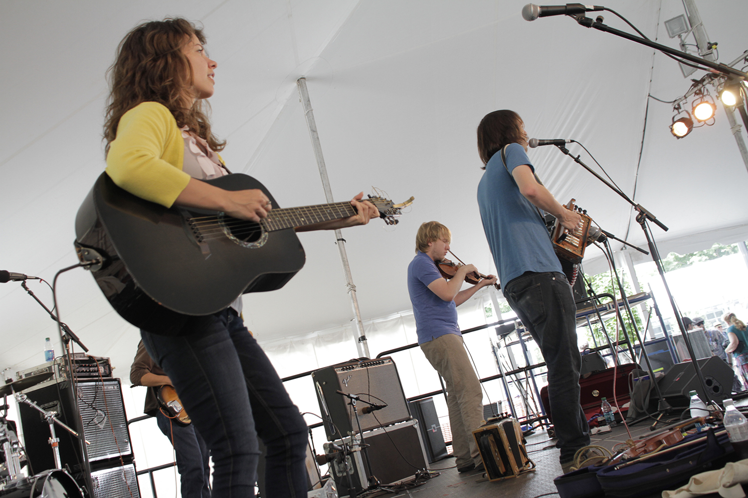 The band plays onstage under an outdoor tent, with their instruments which are: guitars, fiddle, and accordion.