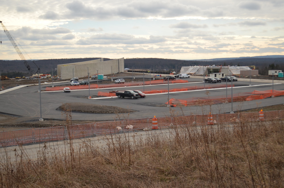 View of gray concrete building, parking area, rectangular building encased in pastic sheeting, and construction equipment