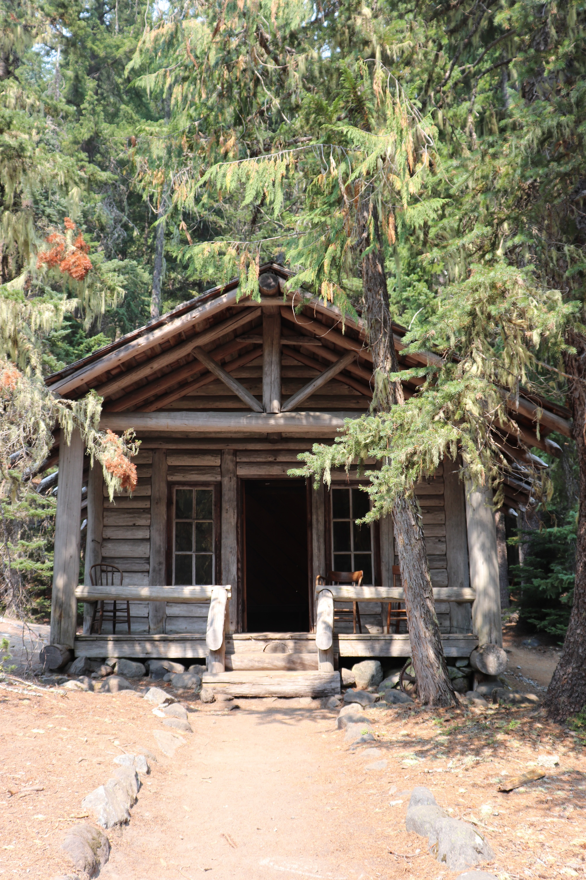 The front of a log cabin with a covered log porch and two windows on either side of the front door. A rock-lined path leads to the porch. 