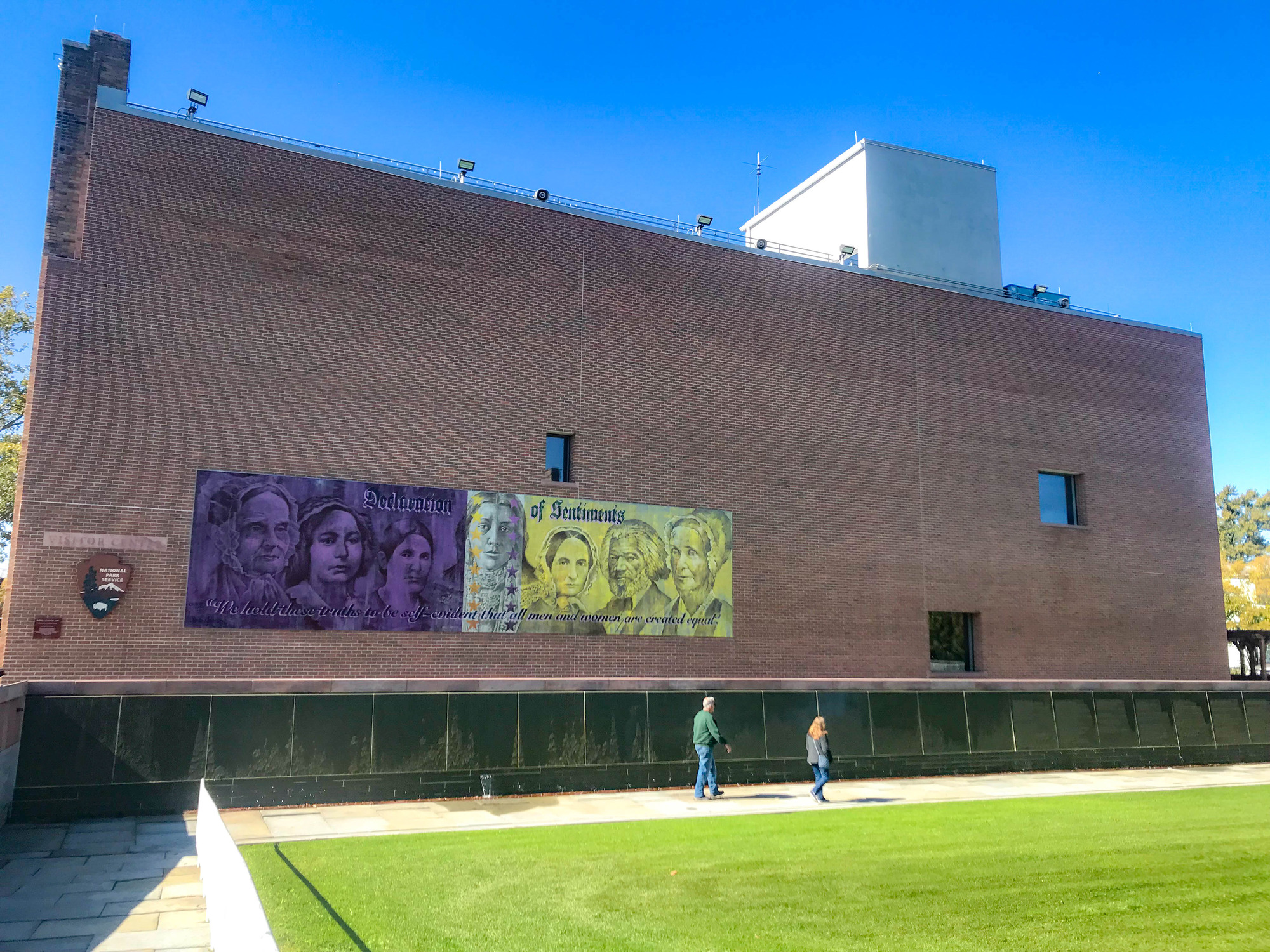 Visitors walking along a wall outside a brick building with the NPS arrowhead logo and a banner depicting people from the women suffrage movement hanging on the side